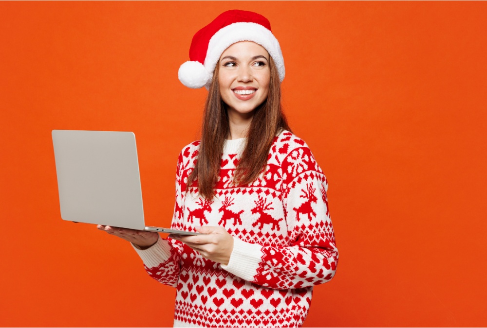 Happy young woman in festive reindeer sweater with laptop on orange background
