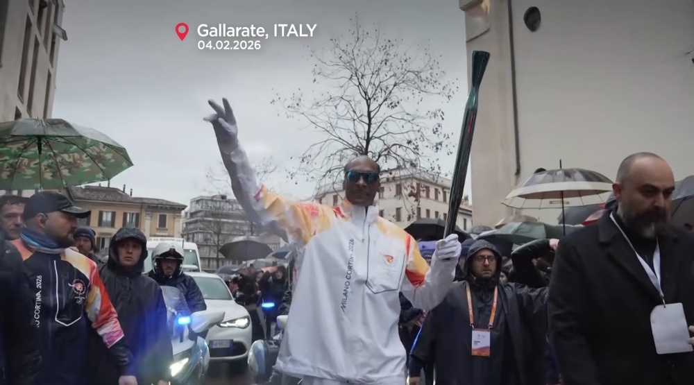 Snoop Dog in white torchbearer suit raises Olympic torch in rainy Gallarate crowd, Italy 2026